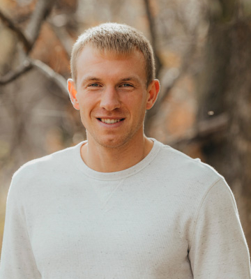 Portrait of a young man with short blond hair wearing a light gray sweater, standing outdoors in a wooded area with fall colors in the background.