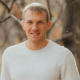 Portrait of a young man with short blond hair wearing a light gray sweater, standing outdoors in a wooded area with fall colors in the background.