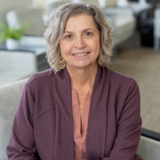 Smiling older woman with short gray hair wearing a purple cardigan, seated in a bright living room.