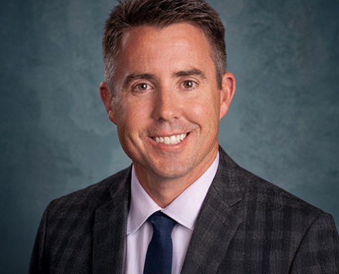 Professional headshot: smiling man in a dark plaid suit with a navy tie, against a teal backdrop.