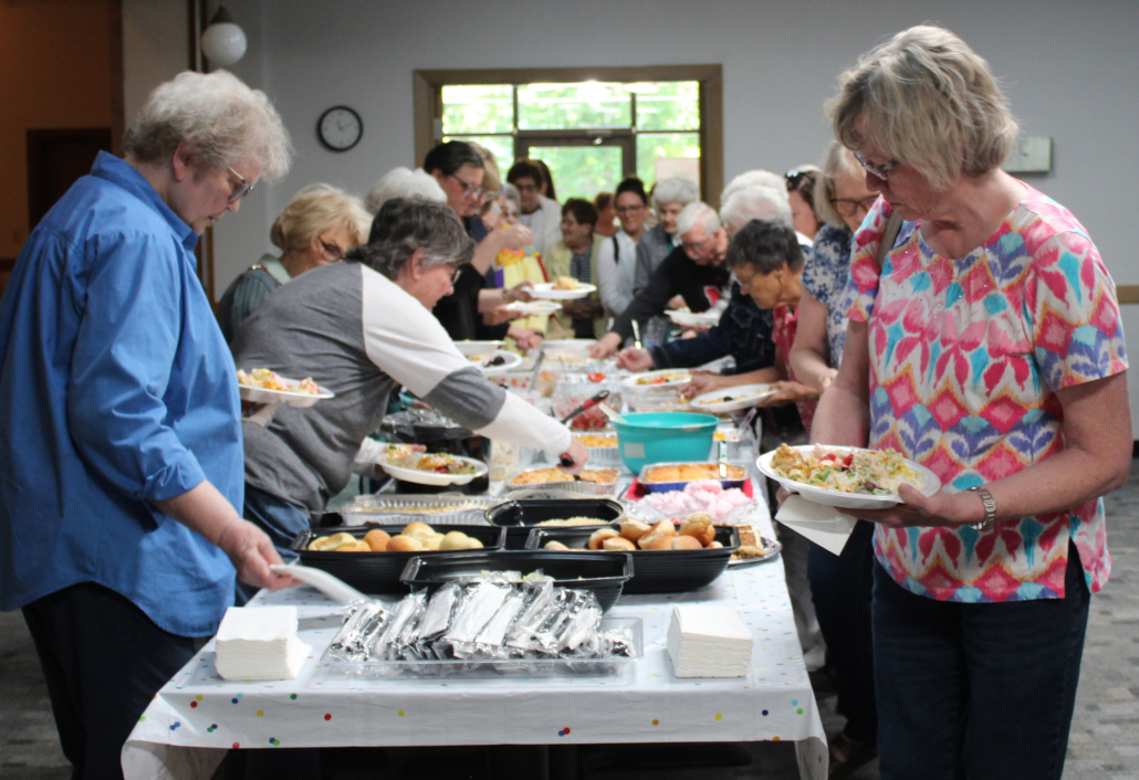 People line up at a buffet, serving themselves from a long table of assorted dishes at a community event.