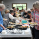 People line up at a buffet, serving themselves from a long table of assorted dishes at a community event.