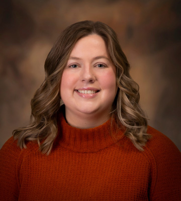 Portrait of a smiling woman with wavy light brown hair wearing an orange knit sweater against a warm brown studio backdrop