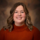 Portrait of a smiling woman with wavy light brown hair wearing an orange knit sweater against a warm brown studio backdrop
