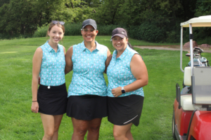 Three women in matching light-blue patterned tops and black bottoms pose together on a grassy golf course beside a golf cart.