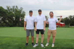 Three men in white polo shirts stand on a golf course green, posing with clubs and a flag in the background.